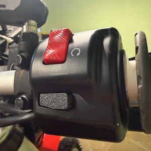 Op de afbeelding: Close-up van een motorfiets stuurmodule. De module is zwart met een rode schakelaar en een getextureerde knop. Een wit pijlsymbool is zichtbaar op de module.