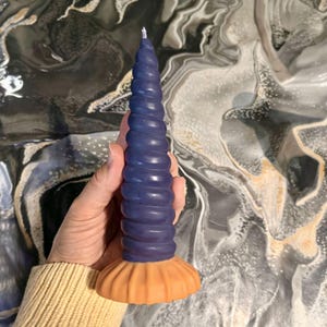 Può includere: Una candela unica a forma di spirale nei toni del blu e dell'arancione. La candela ha un design strutturato e scanalato e una base ampia e scanalata. La parte superiore della candela è blu scuro, che sfuma in un arancione più chiaro alla base. La candela è tenuta in mano.