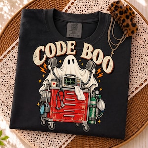 Puede incluir: Una camiseta negra con un gráfico de un fantasma blanco detrás de un carro médico rojo. El texto CODE BOO está impreso en letras grandes de color beige sobre el fantasma. El carro incluye un monitor, un estetoscopio y un tanque de oxígeno.