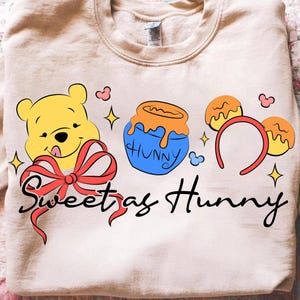 May include: A beige sweatshirt with a cartoon design of Winnie the Pooh, a honey pot, and Mickey Mouse ears. The words "Sweet as Hunny" are written in black script. The design includes red, yellow, blue, and orange accents.