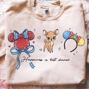 May include: A light beige sweatshirt featuring cartoon graphics of a red Mickey Mouse head with a blue bow, a fawn, and a Minnie Mouse ear headband. The text "Happiness is best shared" is written in black script.