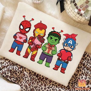 May include: A cream-colored pillow featuring cartoon versions of Spider-Man, Iron Man, Hulk, and Captain America, each holding Valentine's Day gifts. The characters are in their signature colors, with heart-shaped antennae.