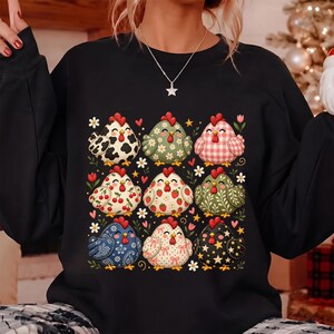 K&ouml;nnte beinhalten: Ein schwarzes Langarm-Sweatshirt mit einem Grafikdruck von neun stilisierten H&uuml;hnern in einem Raster. Jedes Huhn zeigt ein einzigartiges Muster, darunter Kuhflecken, Blumen, Vichy-Karo, Kirschen, Erdbeeren, Paisley, Punkte und Sternmotive.