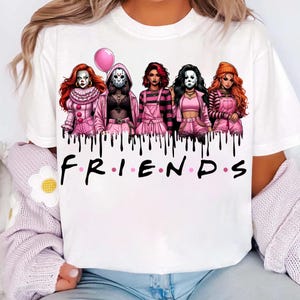 May include: White t-shirt featuring a graphic of five characters in pink outfits, each inspired by horror movie villains. The word "FRIENDS" is printed in black below the characters, with a pink balloon above the "I".