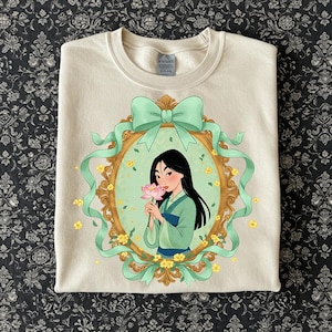K&ouml;nnte beinhalten: Cremefarbenes Sweatshirt mit einer gerahmten Illustration einer Frau in einem gr&uuml;nen und blauen Outfit, die eine rosa Blume h&auml;lt. Der Rahmen ist mit einer gr&uuml;nen Schleife und B&auml;ndern verziert, umgeben von gelben Blumen. Das Sweatshirt hat einen Rundhalsausschnitt.