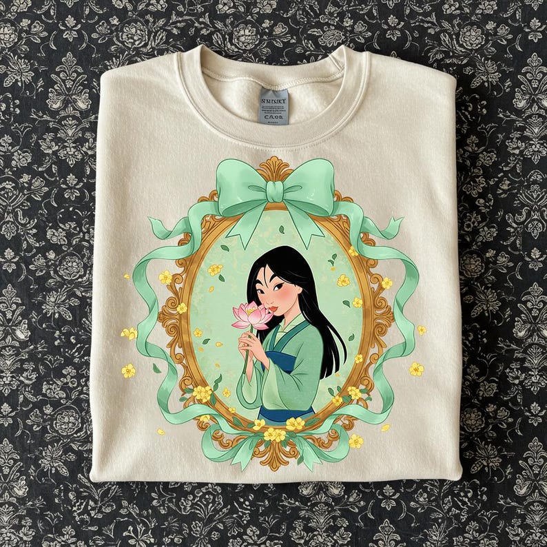 K&ouml;nnte beinhalten: Cremefarbenes Sweatshirt mit einer gerahmten Illustration einer Frau in einem gr&uuml;nen und blauen Outfit, die eine rosa Blume h&auml;lt. Der Rahmen ist mit einer gr&uuml;nen Schleife und B&auml;ndern verziert, umgeben von gelben Blumen. Das Sweatshirt hat einen Rundhalsausschnitt.