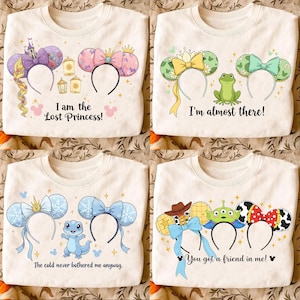 Op de afbeelding: Vier witte sweatshirts met thematische muisoren-hoofdbanden en tekst. Ontwerpen omvatten een paars Rapunzel-thema, een roze strik, groene oren met een kikker en blauwe oren met een hagedis. Tekst bevat "I am the Lost Princess!" en "You got a friend in me!"