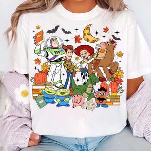 May include: White t-shirt with a vibrant graphic of Toy Story characters, such as Buzz Lightyear and Woody, set against an autumnal backdrop with pumpkins and leaves. The design also features a crescent moon and bats.