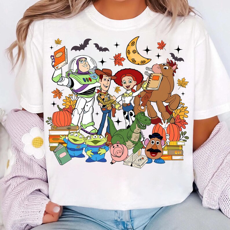May include: White t-shirt with a vibrant graphic of Toy Story characters, such as Buzz Lightyear and Woody, set against an autumnal backdrop with pumpkins and leaves. The design also features a crescent moon and bats.