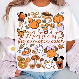 Könnte beinhalten: Weißes T-Shirt mit einem herbstlichen Motiv. Das Design umfasst orangefarbene Kürbisse, Elemente in Mickey-Mouse-Form, Herbstleckereien und den Text "Meet me at the pumpkin patch" in brauner Schrift. Ein lässiges, saisonales Kleidungsstück.