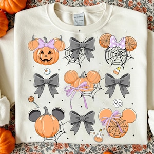 May include: Cream-colored sweatshirt featuring Halloween-themed graphics. The design includes orange pumpkins with mouse ears, gray bows, spiderwebs, and a candy corn. The graphics are arranged in a repeating pattern across the front of the shirt.