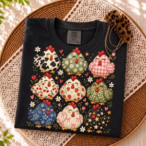 K&ouml;nnte beinhalten: Ein schwarzes Kurzarm-T-Shirt mit einem Grafikdruck von neun stilisierten H&uuml;hnern mit verschiedenen Mustern, darunter Kuhflecken, Vichy-Karo, Kirschen, Erdbeeren und Sterne. Das T-Shirt hat ein Comfort Colors Markenetikett am inneren Halsausschnitt.