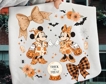 Könnte beinhalten: Ein weißes Halloween-Handtuch mit zwei Zeichentrickmäusen in orangefarbenen Patchwork-Kostümen, die Kürbis-Süßigkeitenkörbe halten. Das Design umfasst orangefarbene Schleifen, Blumen, Fledermäuse und den Text "TRICK OR TREAT" in einer Kristallkugel.