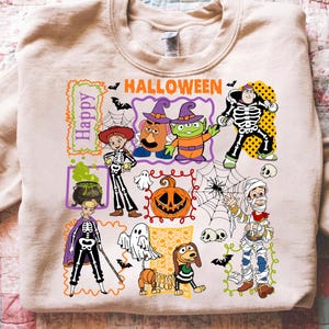 May include: Beige Halloween-themed sweatshirt with cartoon characters in costumes. The design includes a pumpkin, ghosts, and the words "Happy Halloween". The sweatshirt is made of a soft material.