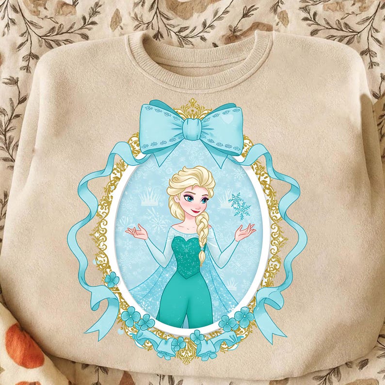 K&ouml;nnte beinhalten: Beiges Sweatshirt mit hellblauer Schleife und ovalem Rahmen. Der Rahmen zeigt eine Cartoon-Zeichnung einer blonden Frau in einem t&uuml;rkisfarbenen Kleid. Der Rahmen hat einen goldenen Rand und ist von hellblauen B&auml;ndern und floralen Akzenten umgeben.