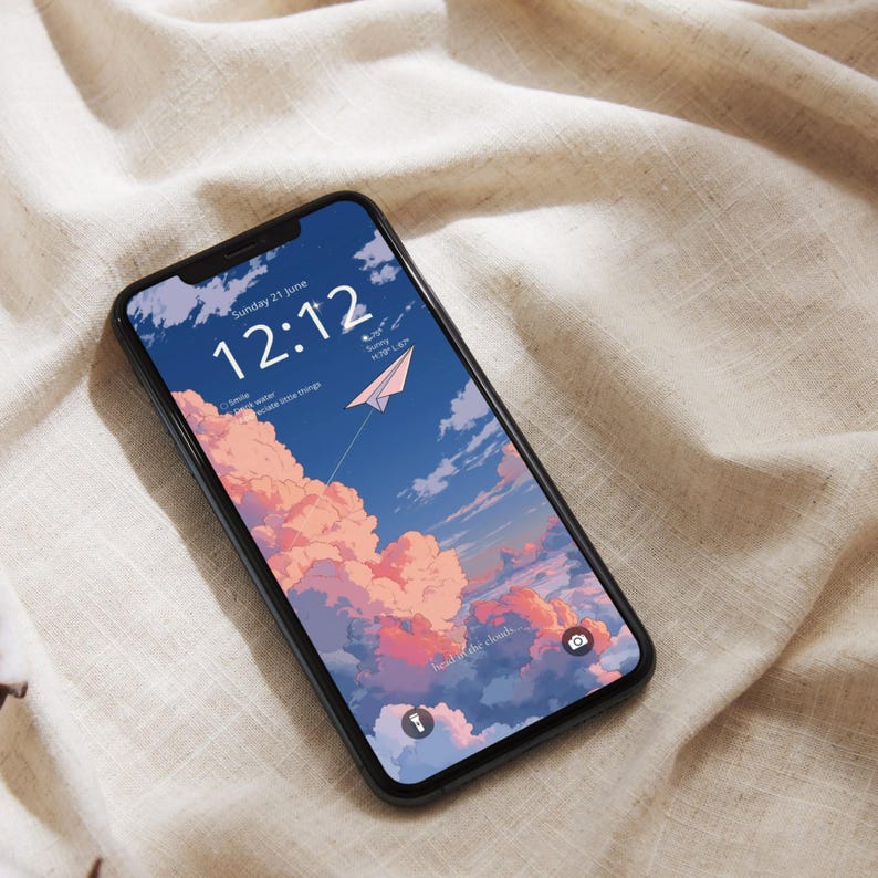 May include: A black smartphone displays a wallpaper with a sky-themed illustration. The image features a blue sky with pink clouds, a paper airplane, and the time 12:12. The phone rests on a beige fabric surface.