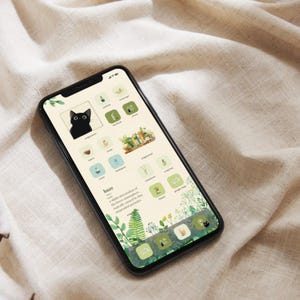 May include: A black smartphone displays a custom home screen with a cat icon, botanical illustrations, and app icons in green and beige. The screen features the word "haze" and a definition. The phone is set against a neutral fabric background.