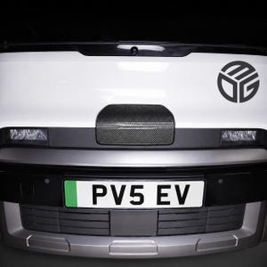 May include: The front of a white vehicle featuring a black circular logo with the letters MDG. A carbon fibre textured panel is centred above the bumper. The number plate displays the text PV5 EV with a green section on the left side.