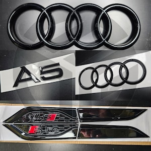 May include: Black car emblems and badges, including the Audi rings logo, A5 emblem, and S-Line badges. The emblems are glossy black with red accents on the S-Line badges. The image is taken from a top-down perspective.