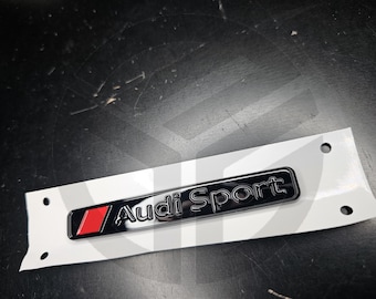 May include: Black and chrome Audi Sport emblem with a red accent, mounted on a white backing with three holes. The emblem features the Audi Sport logo in a sleek, modern font.