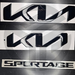 Puede incluir: Tres emblemas de coche negros y plateados. Dos presentan las letras estilizadas "KI" en un acabado negro brillante. El tercer emblema dice "SPORTAGE" en letras plateadas.