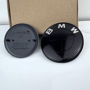 May include: Two black and silver BMW emblem badges. One badge features the BMW logo with the letters "B", "M", and "W" in white. The other badge has text and numbers including "DE" and "103334-10".