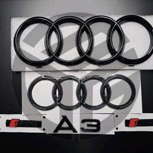Op de afbeelding: Zwart Audi-embleem met vier in elkaar grijpende ringen en de tekst "A3" in zwart. Het embleem bevindt zich op een witte achtergrond.