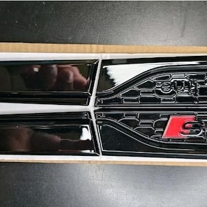 Pode incluir: Quatro emblemas de carro pretos, dois com um design elegante e curvo e dois com um padrão de favo de mel. Os emblemas com o padrão de favo de mel têm o texto "S line" em vermelho. Os emblemas estão sobre uma superfície branca.