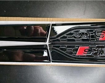 May include: Four black car emblems, two with a sleek, curved design and two with a honeycomb pattern. The emblems with the honeycomb pattern have the text "S line" in red. The emblems are on a white surface.