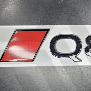 以下が含まれることがあります： 黒と赤のAudi Q8ロゴがついた白い長方形のステッカー。"Q8"は黒で、Audiのロゴは赤です。