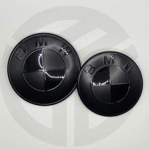 May include: Two black BMW emblems of different sizes. Each emblem features the BMW logo, a circular design with a black outer ring and a divided inner section in black and silver. The BMW letters are visible.