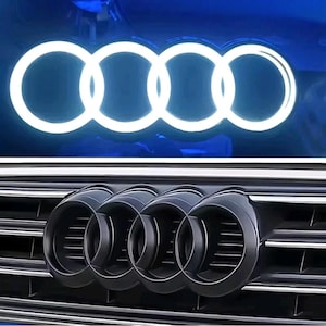 May include: A vehicle emblem featuring four interlocking rings. The top portion displays the rings illuminated in a bright white glow against a dark blue background. The bottom portion shows the matte black emblem mounted on a horizontal grille.