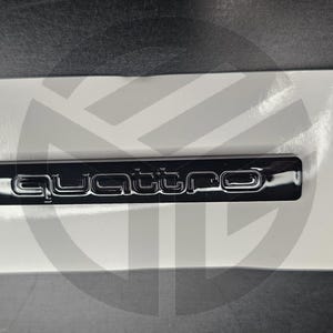 Puede incluir: Un emblema Quattro negro brillante, montado sobre una superficie blanca. La palabra "Quattro" se muestra en una fuente estilizada. El emblema es rectangular y parece ser un accesorio para automóviles.