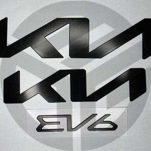 May include: Black Kia EV6 emblem set. The image shows two large Kia logos and a smaller EV6 emblem. The logos are in a modern, angular style, and the EV6 emblem is in a rectangular frame. The background is a light grey with a circular shadow.