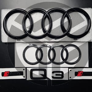 Op de afbeelding: Zwart Audi-ringembleem met vier in elkaar grijpende ringen, en een zwart Audi Q3-embleem met rode S-letters.