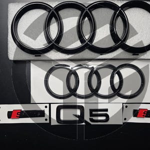 以下が含まれることがあります： 黒色のアウディのエンブレムで、4つの輪が絡み合い、黒色の「Q5」の文字が書かれています。エンブレムは白い背景の上にあります。