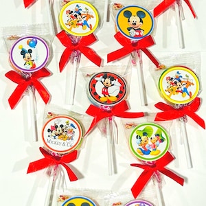 May include: Lollipops featuring various images of Mickey Mouse and friends, each lollipop is individually wrapped and tied with a red ribbon. The lollipops have a white stick and are arranged on a white surface.