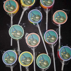 May include: A collection of wrapped lollipops with a cartoon zombie wearing a party hat. The lollipops have a rainbow-colored candy base and a white stick. The zombie image is in a blue circle with a yellow border.