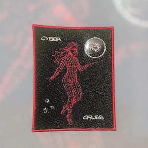 Puede incluir: Un parche bordado rectangular con fondo negro y borde rojo. Presenta una silueta de alambre roja de una persona, el texto CYBER, el texto CRUES, tres pequeñas estrellas blancas y un elemento circular con un borde blanco.