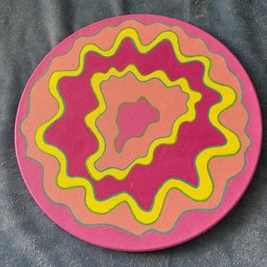 May include: A round, pink canvas painting with concentric circles of yellow, orange, and green wavy lines.