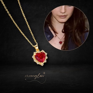 Puede incluir: Un collar de cadena dorada con un colgante en forma de corazón rojo. El colgante está adornado con piedras brillantes transparentes alrededor del borde.