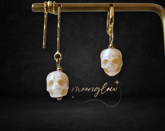 May include: A pair of gold skull earrings with white pearl skull charms. The earrings are hanging from a gold stand.
