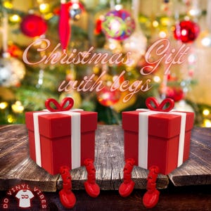 May include: Two red gift boxes with white ribbon and bow accents, each with red legs, sit on a wooden surface. The text "Christmas Gift with legs" is above the gifts. A Christmas tree with ornaments is in the background.