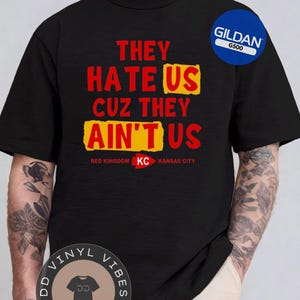 Op de afbeelding: Zwart t-shirt met de tekst "THEY HATE US CUZ THEY AIN'T US" in rood en geel. Het shirt heeft ook "RED KINGDOM KC KANSAS CITY". Een blauw Gildan G500-label is zichtbaar. Onderaan staat een DD Vinyl Vibes-logo.