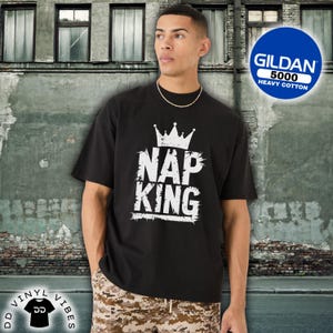 Könnte beinhalten: Schwarzes T-Shirt mit dem weißen Text "NAP KING" und einer Kronengrafik. Das T-Shirt besteht aus schwerer Baumwolle, einem Gildan 5000. Das Model trägt eine goldene Kette. Der Hintergrund ist eine verwitterte Backsteinmauer.