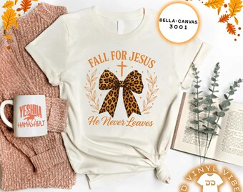 May include: Off-white t-shirt featuring the text "Fall for Jesus, He Never Leaves" and a leopard print bow design. A white mug with the word "Yeshua" is on the left. A book and a sweater are also in the image.