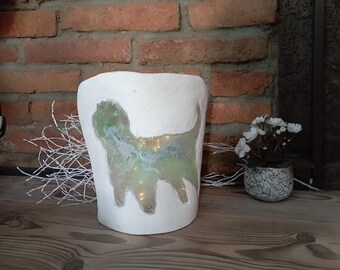 May include: White ceramic lamp with a green silhouette of a dog inside. The lamp is sitting on a wooden surface with white branches in the background.