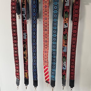 May include: A collection of colorful, patterned camera straps hanging on a brass rod. The straps feature intricate designs in red, black, blue, and green, with some displaying text. Each strap has metal clips for attachment.
