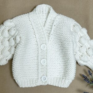 May include: A white, hand-knitted cardigan with a V-neck and three large buttons down the front. The sleeves feature a textured, bubble-like design. The soft, off-white color and cozy knit suggest a baby or toddler garment.