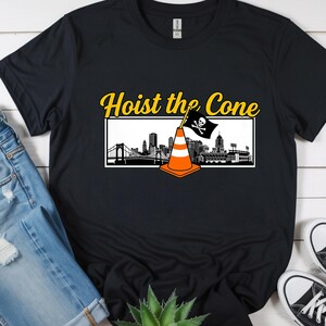 May include: Black t-shirt with the text "Hoist the Cone" in yellow, above a graphic of a traffic cone with a pirate flag and a city skyline. The shirt is paired with blue jeans and black sneakers.